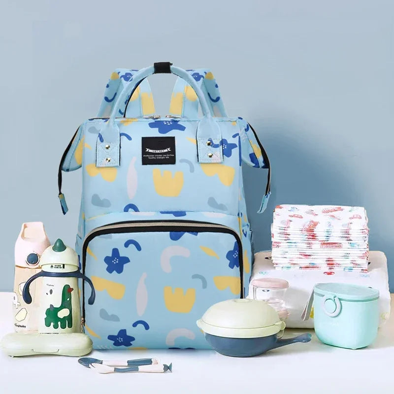 Sac a langer entoure de biberons, couches et boites de lait montrant sa grande capacite pour organiser tout le necessaire de bebe en une seule sortie.