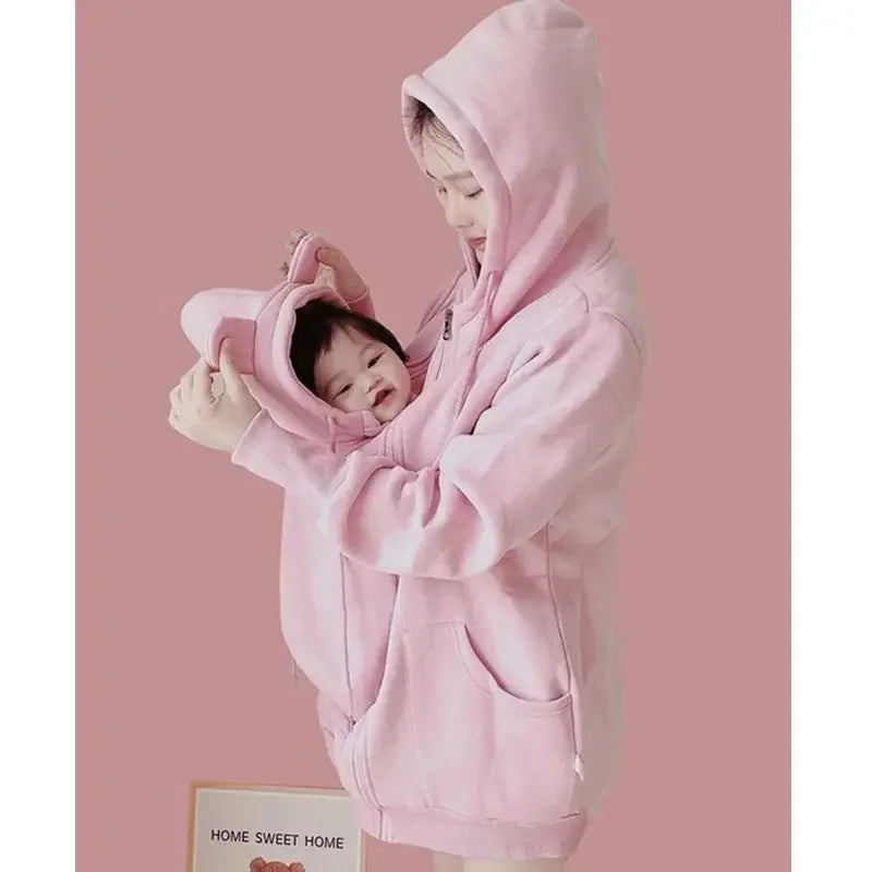 Maman installe son bebe souriant dans la poche de portage du manteau rose, un geste simple pour le garder au chaud.