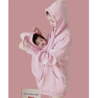 Maman installe son bebe souriant dans la poche de portage du manteau rose, un geste simple pour le garder au chaud.