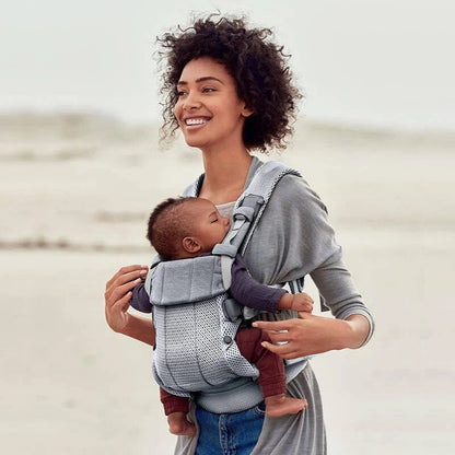 Maman portant son nourrisson endormi en porte bebe physio a la plage, mains libres pour profiter de la balade.