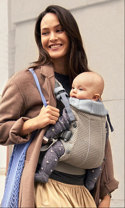 Maman souriante portant son bebe en porte bebe physio lors d'une sortie urbaine, illustrant ergonomie et respirabilite.