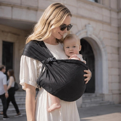 Maman elegante se promenant en ville avec son bebe eveille dans un sling noir bien maintenu contre elle.