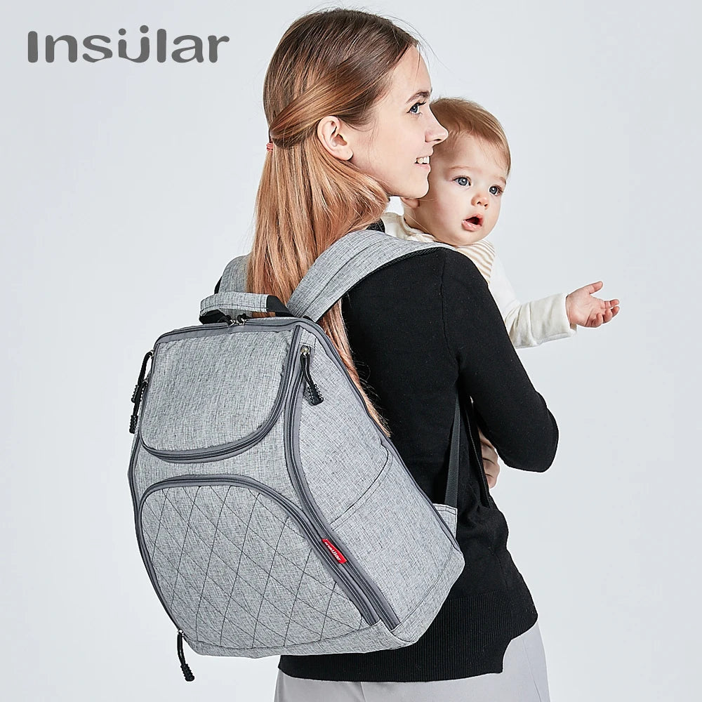 Maman portant le sac a langer a dos tout en tenant son bebe, illustrant la liberte de mouvement au quotidien.