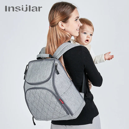 Maman portant le sac a langer a dos tout en tenant son bebe, illustrant la liberte de mouvement au quotidien.