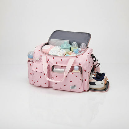 Sac a langer rose motif fraises presente en trois coloris avec grand compartiment ouvert rempli d'affaires de bebe et espace lateral dedie aux chaussures.