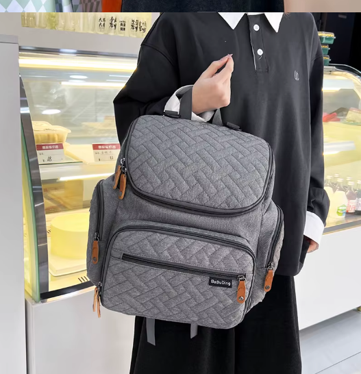 Sac a langer gris porte a la main en ville, illustrant sa polyvalence entre sac a dos et sac de tous les jours.